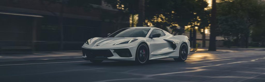 White Chevrolet Corvette driving on a city street at sunset.